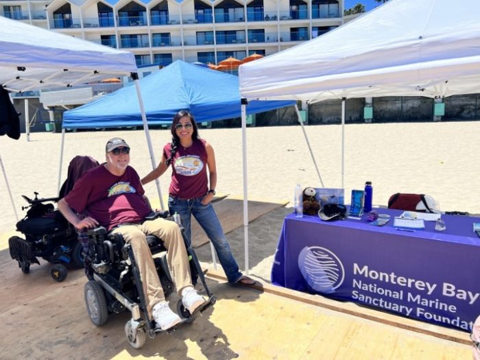 (Image description: An image of Foster Anderson, founder of Shared Adventures, and Elaine Reyes, volunteer and representative of the Monterey Bay National Marine Sanctuary Foundation standing next to a booth. They are both smiling.) 